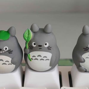 May include: Three gray Totoro-themed keycaps on a white keyboard. One keycap has a green leaf, another has a green leaf hat. The keycaps are on the 4 and 5 keys. The keycaps are a cute and whimsical addition to any keyboard.