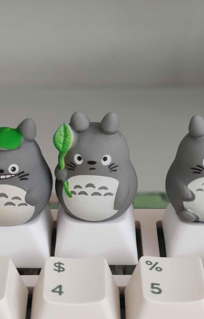 May include: Three gray Totoro-themed keycaps on a white keyboard. One keycap has a green leaf, another has a green leaf hat. The keycaps are on the 4 and 5 keys. The keycaps are a cute and whimsical addition to any keyboard.
