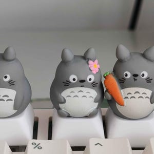 May include: Three gray Totoro-inspired keycaps on a white keyboard. One has a pink flower, and another holds a small orange carrot. The keycaps have white bellies and black eyes, adding a touch of whimsy to any keyboard setup.