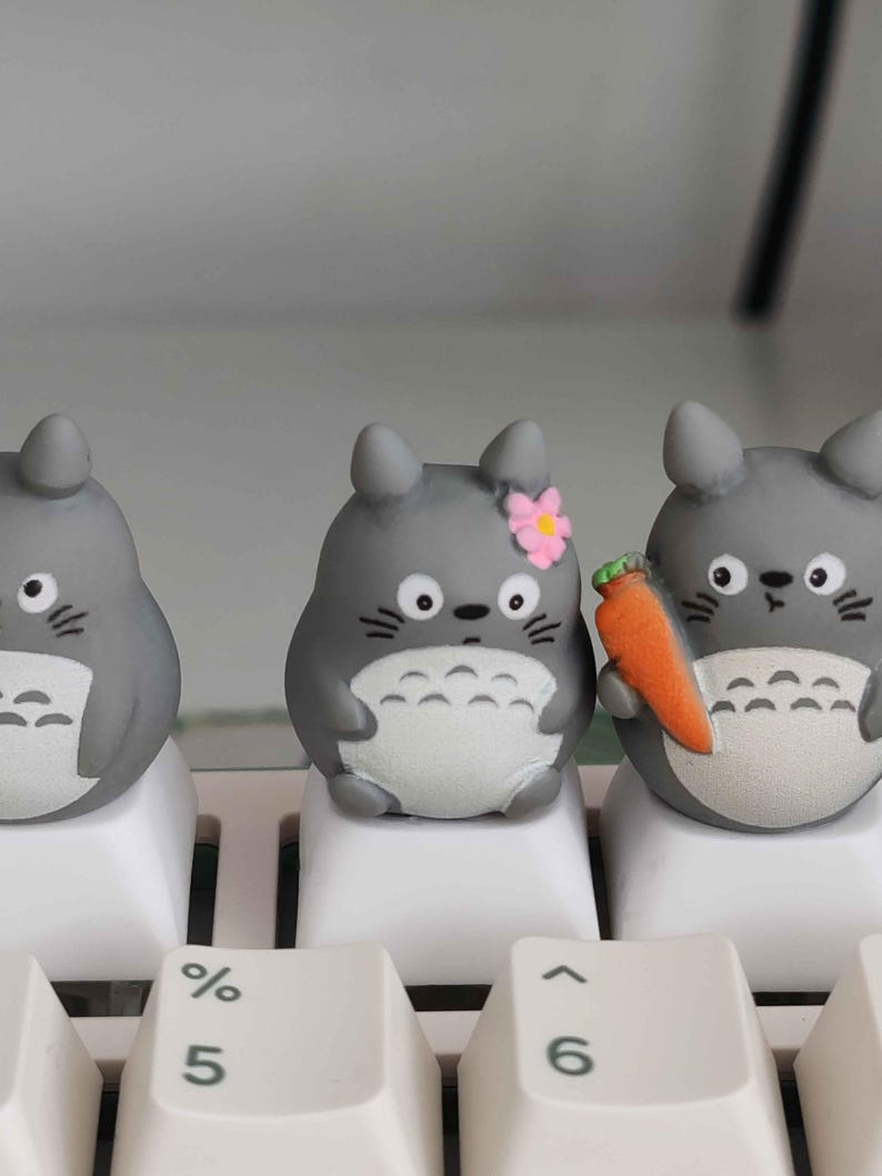 May include: Three gray Totoro-inspired keycaps on a white keyboard. One has a pink flower, and another holds a small orange carrot. The keycaps have white bellies and black eyes, adding a touch of whimsy to any keyboard setup.