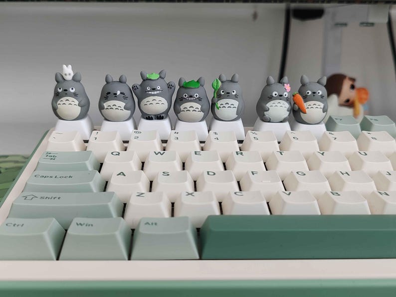 May include: A mechanical keyboard with white keycaps and a green accent. Adorning the top row are seven gray Totoro-themed keycap figures, each with unique accessories. The keyboard's design is clean and modern, with a focus on the custom keycaps.