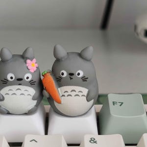 May include: Two gray Totoro-shaped keycaps with black whiskers and white bellies sit on a white keyboard. One holds a carrot, the other a pink flower. A light green F7 key is visible.