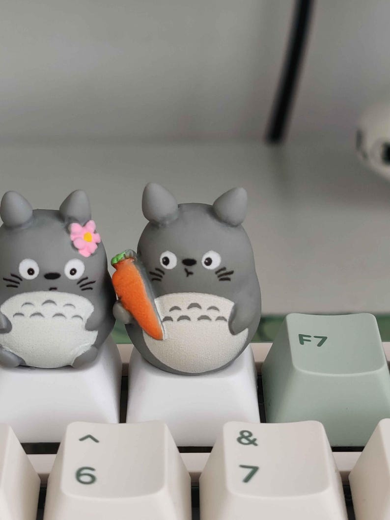 May include: Two gray Totoro-shaped keycaps with black whiskers and white bellies sit on a white keyboard. One holds a carrot, the other a pink flower. A light green F7 key is visible.