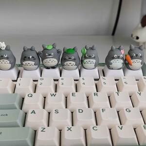 May include: A keyboard with custom keycaps featuring gray Totoro figures. The keycaps are placed on white keys with light green accents. The keyboard has a standard QWERTY layout with numbers and symbols.