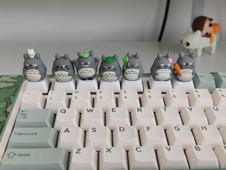 May include: A keyboard with custom keycaps featuring gray Totoro figures. The keycaps are placed on white keys with light green accents. The keyboard has a standard QWERTY layout with numbers and symbols.