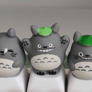 May include: Three gray Totoro-inspired keycaps with varying expressions. The central keycap has a wide smile and raised arms, while the others have different facial expressions. Each keycap has a green leaf on its head and is set on white keyboard keys.