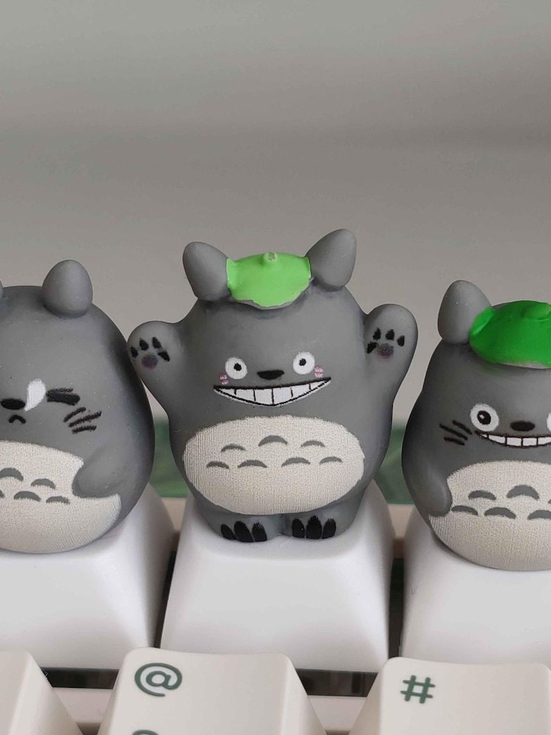 May include: Three gray Totoro-inspired keycaps with varying expressions. The central keycap has a wide smile and raised arms, while the others have different facial expressions. Each keycap has a green leaf on its head and is set on white keyboard keys.