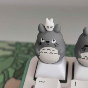 May include: A gray Totoro-themed keycap with a small white creature on top, sitting on a white keyboard key. The keycap has black eyes and a white belly. The keyboard has a mint green border.