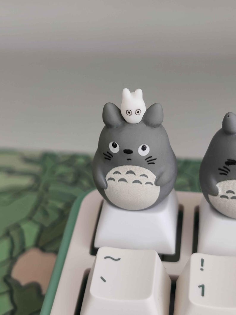 May include: A gray Totoro-themed keycap with a small white creature on top, sitting on a white keyboard key. The keycap has black eyes and a white belly. The keyboard has a mint green border.