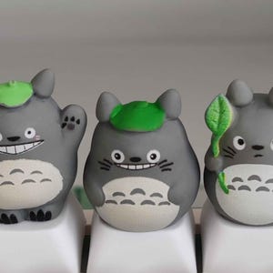 May include: Three gray Totoro figurines with green leaf hats and white bellies sit on white keyboard keys. One figure waves, another holds a leaf, and all have smiling faces. The keyboard keys have the numbers 3 and 4, and the symbols # and $.