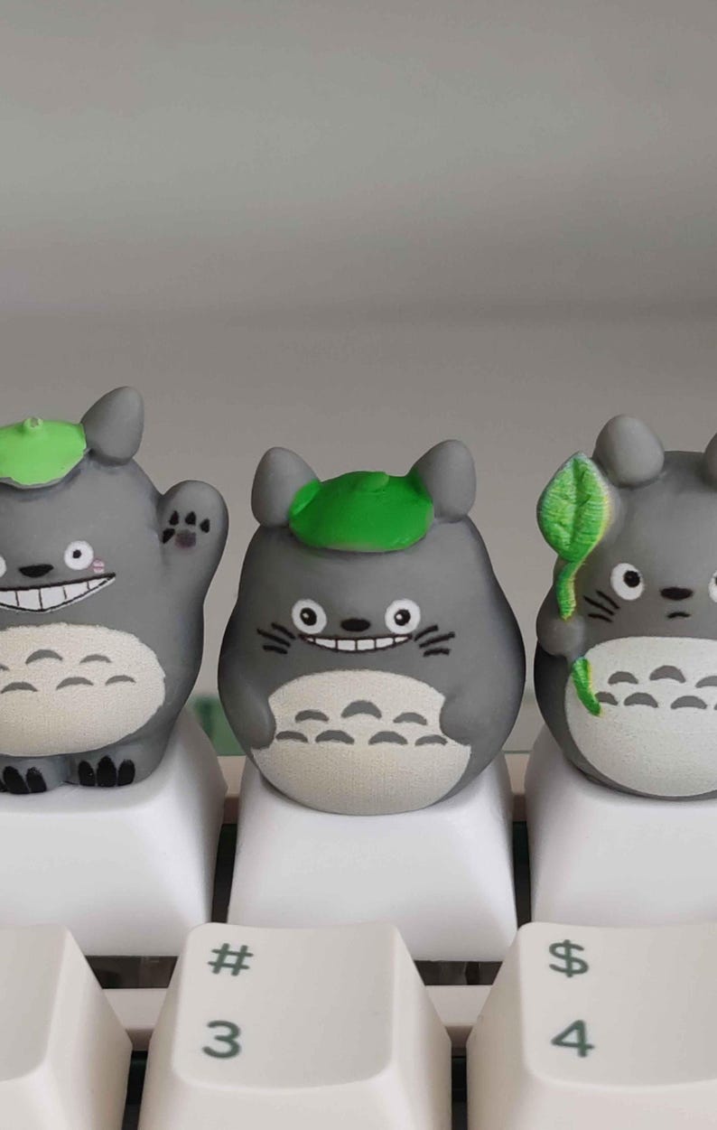 May include: Three gray Totoro figurines with green leaf hats and white bellies sit on white keyboard keys. One figure waves, another holds a leaf, and all have smiling faces. The keyboard keys have the numbers 3 and 4, and the symbols # and $.