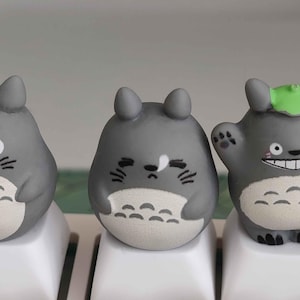 May include: Three gray Totoro-themed keycaps on a white keyboard. The keycaps have a rounded shape with black facial details and white bellies. One Totoro has a green leaf on its head.
