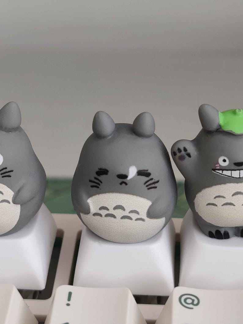 May include: Three gray Totoro-themed keycaps on a white keyboard. The keycaps have a rounded shape with black facial details and white bellies. One Totoro has a green leaf on its head.