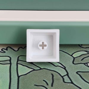 May include: A white square keycap with a cross-shaped stem mount. The keycap is set against a background of green and white panels and a green patterned surface.