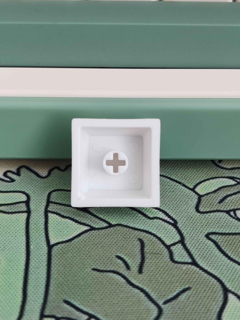 May include: A white square keycap with a cross-shaped stem mount. The keycap is set against a background of green and white panels and a green patterned surface.