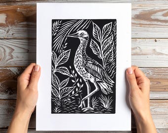 May include: A black and white linocut print of a bird with long legs and a long beak, surrounded by stylized foliage. The artwork is displayed in a white mat, held by hands against a wooden background. The bird is the focal point.