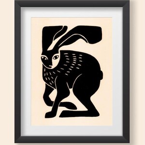 May include: A framed print featuring a stylized black silhouette of a hare on a cream-colored background. The hare is depicted in a seated position with long ears, and the artwork is enclosed within a simple, thin black rectangular frame.