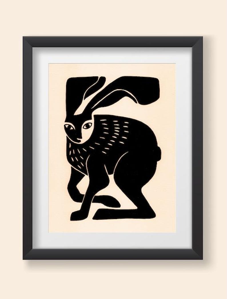 May include: A framed print featuring a stylized black silhouette of a hare on a cream-colored background. The hare is depicted in a seated position with long ears, and the artwork is enclosed within a simple, thin black rectangular frame.