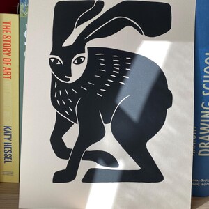 May include: A black and white print on off-white paper featuring a stylized graphic illustration of a hare. The hare is depicted in a seated position with long ears, almond-shaped eyes, and short, dash-like markings on its body and ears.