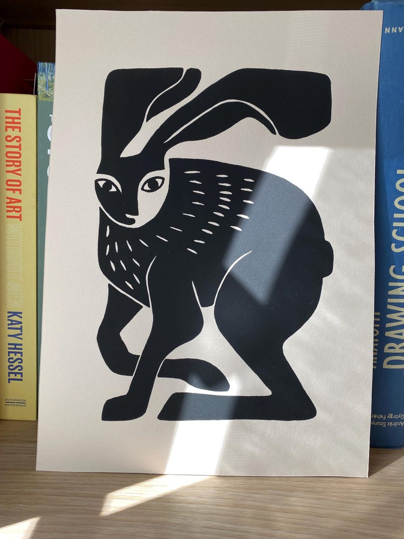 May include: A black and white print on off-white paper featuring a stylized graphic illustration of a hare. The hare is depicted in a seated position with long ears, almond-shaped eyes, and short, dash-like markings on its body and ears.