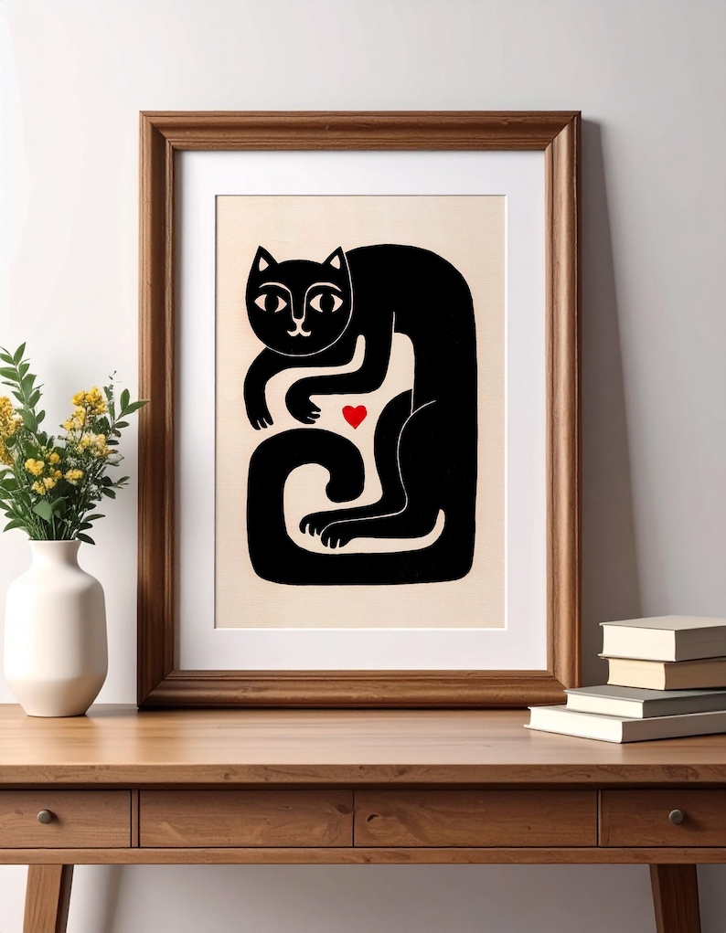 May include: A framed art print featuring a stylized black cat illustration on a cream background. The cat is depicted in a curled, seated position with a small, solid red heart centered between its front paws and its long, curved tail.