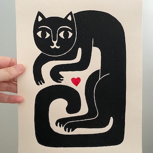 May include: A black ink block print on off-white paper featuring a stylized cat with a long, curved tail. The cat is depicted in a seated position with its front paws tucked inward. A small, solid red heart is positioned between the cat's paws.