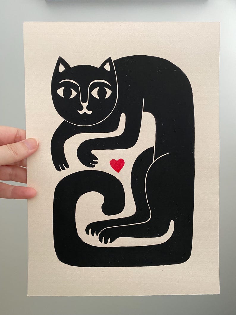 May include: A black ink block print on off-white paper featuring a stylized cat with a long, curved tail. The cat is depicted in a seated position with its front paws tucked inward. A small, solid red heart is positioned between the cat's paws.