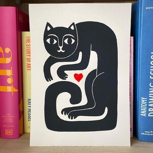 May include: A rectangular print on off-white paper featuring a stylized black cat illustration. The cat is depicted in a seated, curved posture with a small, solid red heart positioned centrally between its front paws and its long, curled tail.
