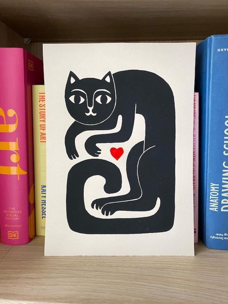 May include: A rectangular print on off-white paper featuring a stylized black cat illustration. The cat is depicted in a seated, curved posture with a small, solid red heart positioned centrally between its front paws and its long, curled tail.