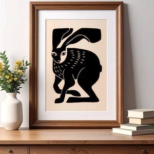 May include: A framed wall art print featuring a stylized black silhouette of a hare on a light beige background. The hare is depicted in a side profile with long ears and textured markings on its chest, enclosed in a simple wooden frame.