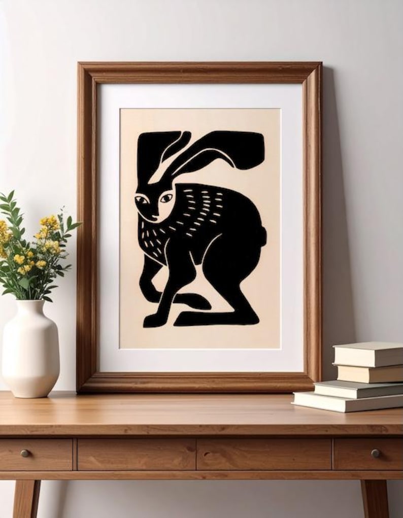 May include: A framed wall art print featuring a stylized black silhouette of a hare on a light beige background. The hare is depicted in a side profile with long ears and textured markings on its chest, enclosed in a simple wooden frame.