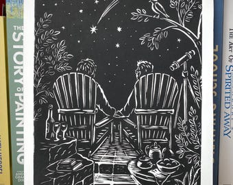 May include: A black and white linocut print depicting two figures sitting in chairs, holding hands while looking at a starry night sky with a shooting star. A bird sits on a tree branch, a telescope stands nearby, and a table holds tea and wine.