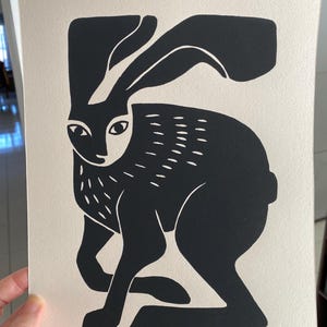May include: A rectangular print on off-white paper featuring a high-contrast black ink illustration of a hare. The hare is depicted in a seated position with long ears, stylized fur markings on the chest, and a solid black body silhouette.