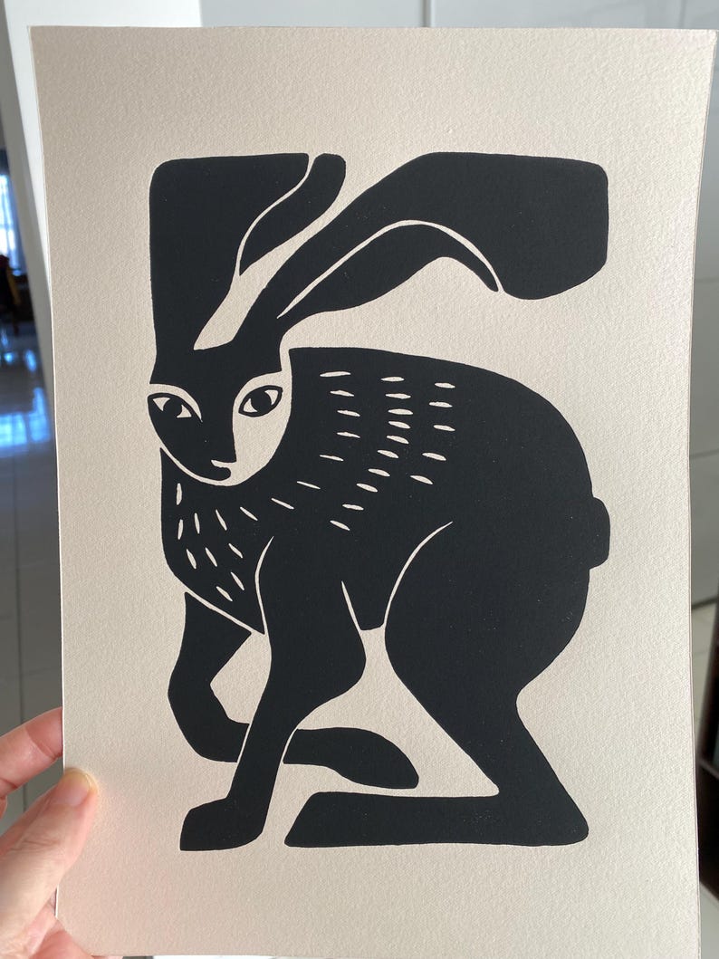 May include: A rectangular print on off-white paper featuring a high-contrast black ink illustration of a hare. The hare is depicted in a seated position with long ears, stylized fur markings on the chest, and a solid black body silhouette.