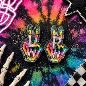 May include: Two acrylic pins shaped like skeleton hands making a peace sign. The hands feature a rainbow gradient pattern with glitter. Each pin has silver-colored metal letters attached to the front, with one reading L MV and the other R MV.