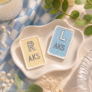May include: Two rectangular decorative tiles with the letters "R" and "L" and the letters "AKS" are displayed on a white, wavy-edged dish. The "R" tile has a yellow background, while the "L" tile has a blue checkered background.