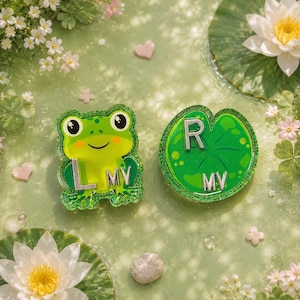 May include: Two glittery acrylic charms, one shaped like a green frog with the silver letters L and MV, and the other shaped like a green lily pad with the silver letters R and MV, both featuring a smooth, glossy finish and vibrant green tones.