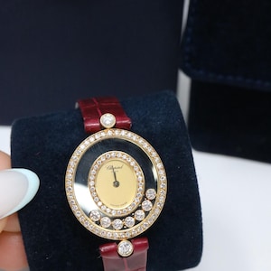 May include: A Chopard wristwatch featuring a round gold case encrusted with diamonds, a gold dial with black hands, and a deep red alligator-textured leather strap. The watch includes floating diamonds within the crystal and a single diamond at each lug.