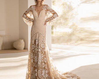 May include: A floor-length gown featuring a deep V-neckline and long, voluminous bishop sleeves. The dress is crafted from a nude-colored base fabric overlaid with intricate, white floral and leaf-patterned embroidery that extends into a train.