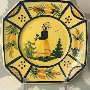 May include: A square ceramic plate with a yellow background featuring a central illustration of a person in a bonnet holding flowers. The border is divided into sections with dark blue lines, decorated with orange floral motifs and small dots.