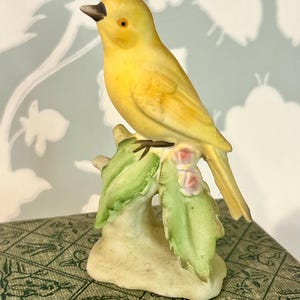 May include: A ceramic figurine of a bright yellow bird perched on a textured branch. The bird has a dark beak and a small eye. The branch features light green leaves and small pink flower buds, resting on a solid, off-white base.