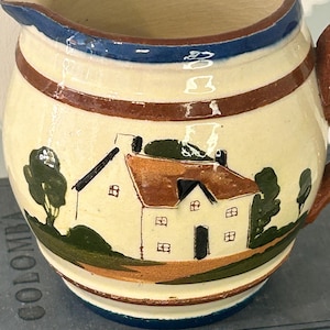 May include: A cream-colored ceramic pitcher featuring a hand-painted scene of a house with a brown roof, green trees, and a path. The rim and base are decorated with horizontal blue and brown bands, and the interior has a crackled finish.