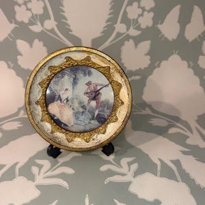 May include: A round decorative plate featuring a central illustration of two figures in period clothing, one playing a lute. The plate has a wide, textured gold-toned border with an ornate, raised star-shaped frame surrounding the central image.