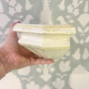 May include: A cream-colored ceramic wall shelf with a glossy finish and a decorative, textured gold-painted edge along the top rim. The shelf features a geometric, angular design with a flat top surface and a tapered, multi-faceted base structure.