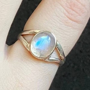 Puede incluir: Un anillo de plata con una piedra de luna ovalada blanca. El anillo tiene un diseño delicado y abierto.