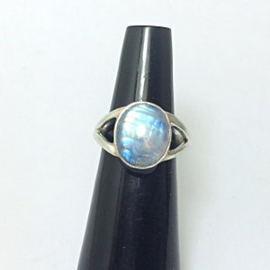 May include: A silver ring featuring a central oval-shaped moonstone cabochon with a translucent, iridescent blue sheen. The metal band has a split-shank design that tapers toward the stone, creating a triangular opening on each side of the setting.