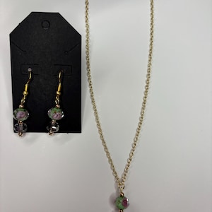 May include: A gold-toned necklace and matching earrings set. The necklace features a chain with a pendant of a green and pink floral bead and a clear crystal bead. The earrings have similar floral beads and clear crystal beads. The jewelry is displayed on a white surface.