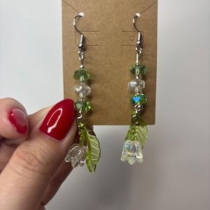 May include: A pair of dangle earrings with silver hooks. Each earring features clear and green beads, a clear flower, and a green leaf. The earrings are displayed on a brown tag against a white background.