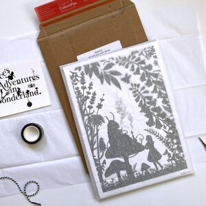 May include: A white card with black text that reads "Alice's Adventures in Wonderland."  A black and white roll of tape, a black and white string, and a white sheet of paper with a gray silhouette of Alice in Wonderland on a mushroom with a caterpillar on top. The silhouette is surrounded by gray leaves and flowers. The paper is wrapped in a white tissue paper.