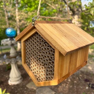 May include: A hexagonal wooden bee house featuring a slanted roof and a front panel filled with numerous small, hollow cardboard tubes. The structure is suspended by a piece of twine attached to a metal hook at the top of the wooden frame.